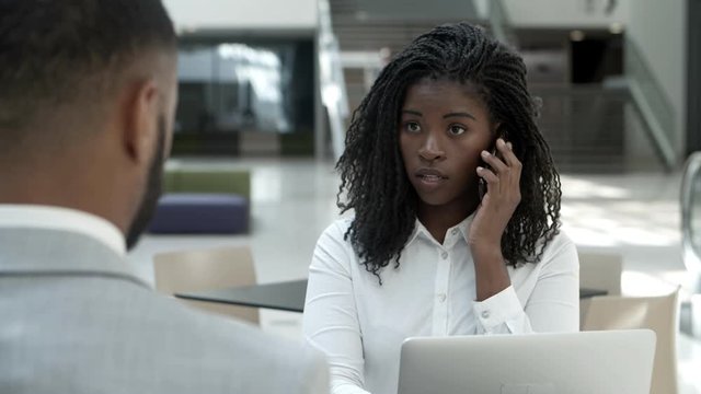 Serious woman talking on smartphone and looking at colleague. Concentrated young businesswoman talking with interlocutor through smartphone while sitting at cafe. Technology concept