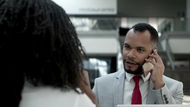 Focused young businessman talking on smartphone. Slow motion shot of African American man communicating with interlocutor while sitting at table with colleague. Business communication concept