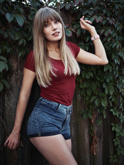 Lifestyle happy blonde woman dressed red blouse blue denim jeans shorts emotionally posing ivy wooden fence european cozy village in evening twilight