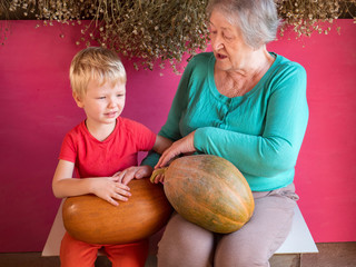 Fototapeta premium elderly woman granny in turquoise sweater with grandson sits with large pumpkin on her lap against background of dry daisy flowers in barn