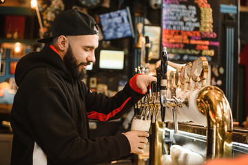 Fototapeta na wymiar smiling barman pouring beer in a bar. Bearded courageous man pours you a foaming drink