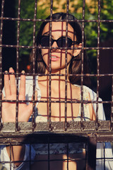 Portrait of girl in sunglasses posing in city behind a trellised fence. Dressed in top with floral print, white shirt, black trousers, waist bag.