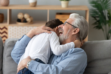 Happy old grandpa embracing little boy grandson cuddling at home
