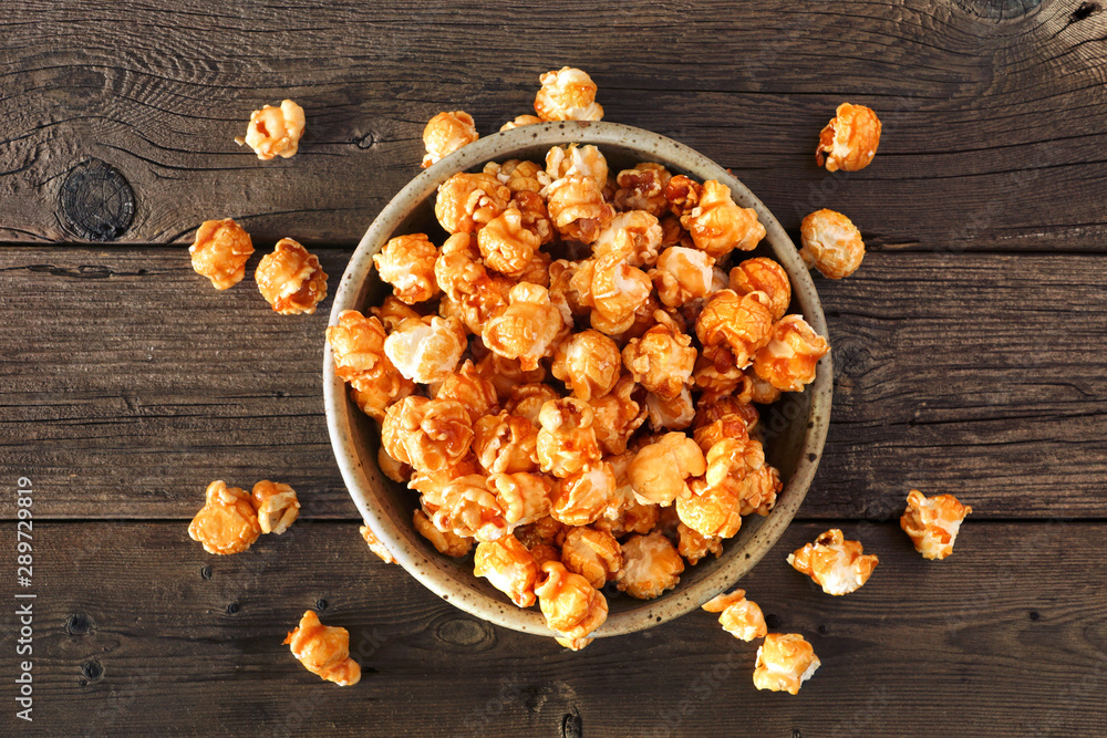 Poster bowl of caramel popcorn with scattered kernels. top view over a rustic wood background.