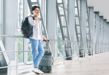 Millennial guy talking on phone, waiting for flight with luggage at airport