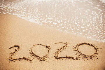 Happy New Year 2020, handwritten on sandy beach. 2020 numbers inscription on golden sand and waves foam at sea beach. Tropical celebration