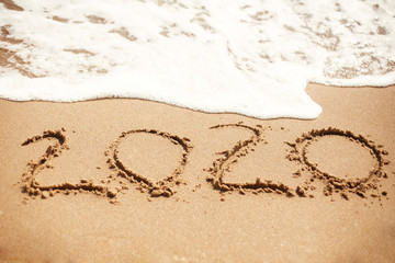 Happy New Year 2020, handwritten on sandy beach. 2020 numbers inscription on golden sand and waves foam at sea beach. Tropical celebration
