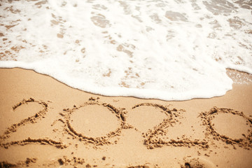 Happy New Year 2020, handwritten on sandy beach. 2020 numbers inscription on golden sand and waves foam at sea beach. Tropical celebration