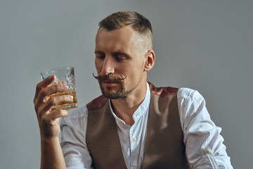 Man with stylish mustache, dressed in classic brown vest, white shirt is sitting at the table, enjoying whiskey. Grey background, close-up shot.