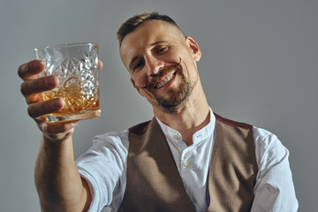 Man with stylish mustache, dressed in classic brown vest, white shirt is sitting at the table, enjoying whiskey. Grey background, close-up shot.