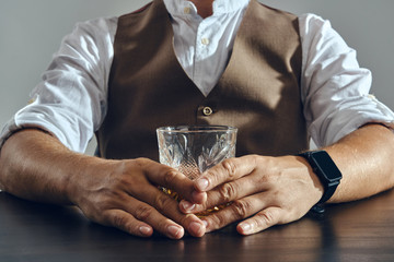 Hands of a man, dressed in a classic brown vest and white shirt is sitting at the table and enjoying whiskey. Grey background, close-up shot.
