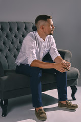 Man with stylish mustache, dressed in white shirt, blue trousers is sitting on dark sofa, holding...