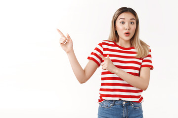 Excited surprised young asian girl blond short hairstyle gasping folding lips amused popping eyes camera as pointing upper left corner wondered questioned talking friend, white background