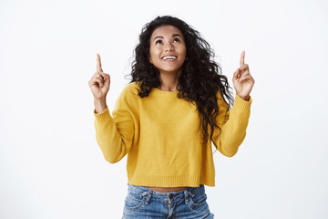 Dreamy cute smiling african american woman in yellow sweater gasping amused, look and pointing fingers up amazed, see awesome opportunity, laughing carefree, white background