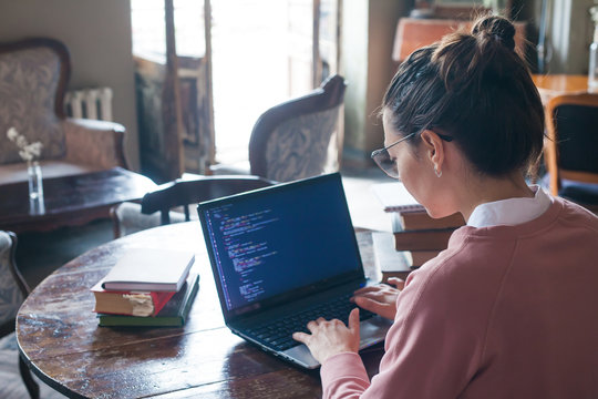 Hacking system. Excited girl with dark hair and in pink sweater and glasses writes code on a laptop in a library.