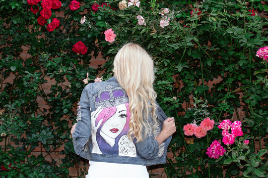 Blonde girl posing in a painted Jean jacket on a background of greenery and flowers.