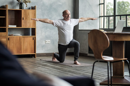 Senior Hispanic Man Standing In Warrior Yoga Pose Variation Practicing In Living Room Alone