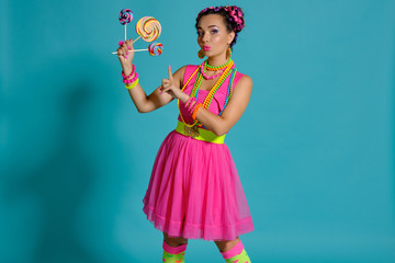 Lovely girl with a multi-colored braids hairstyle and bright make-up, posing in studio against a blue background, holding a lollipop in her hand.