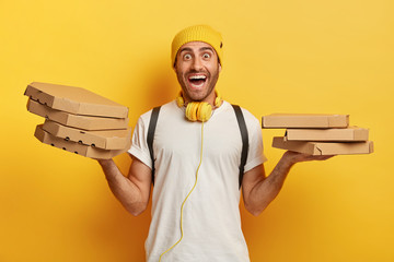 Horizontal shot of happy man holds two piles of carton boxes with pizza, has surprised joyful expression, works as courier in local restaurant, isolated over yellow background, gets many orders