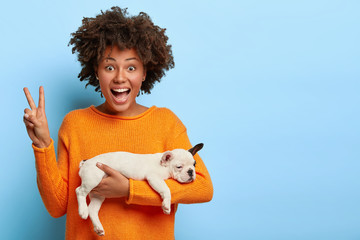 Cheerful Afro American woman makes peace gesture, enjoys good day, carries sleeping puppy, poses...