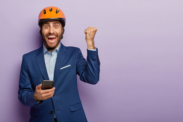 Triumphing happy engineer holds mobile phone, raises clenched fist, uses phone, rejoices being on...