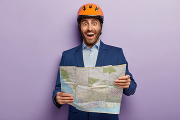 Horizontal shot of happy male architect with map, studies location map where construction site is situated, wears protective helemt, formal wear, isolated on purple wall. Engineer with blueprint
