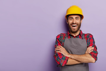 Positive happy foreman in workwear, keeps arms folded, wears yellow hardhat, receives new task from boss, glad to be promoted, stands against purple background, blank free space. Repairman indoor