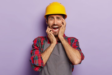 Panicking male architect looks stressfully, worries to have deadline, dressed in special uniform, yellow hardhat, has stubble, poses on purple background. People, occupation, negative emotions concept