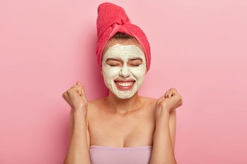 Headshot of beautiful young woman raises clenched fists, applies moisturizing mask on face, has...