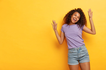 Upbeat cheerful curly haired woman raises arms, feels overjoyed, dances actively, has fun on party, wears casual purple t shirt and jean shorts, poses against yellow studio wall, copy space for text