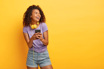 Photo of smiling teenage girl with Afro haircut, uses smartphone for listening music in playlist,...