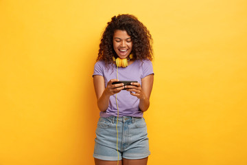 Overjoyed dark skinned female plays smart phone device, being obsessed with online games, spends free time with modern technologies, wears t shirt and shorts, poses against yellow background