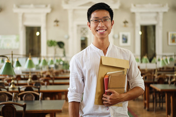 Young smiling asian student in eyeglasses with notepads joyfully looking in camera in library of...