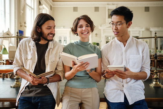 Group of young casual multinational students joyfully studying with book and making notes in library of university