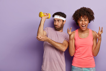 Sport and working on yourself. Unshaven guy raises arm with dumbbell, does exercises for training muscles, irritated curly haired African American woman gestures angrily, dissatisfied by something