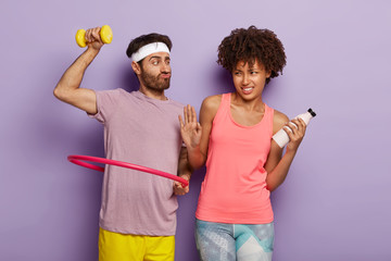 Funny sportsman rotates hula hoop, holds dumbbell, leads healthy lifestyle, displeased Afro...