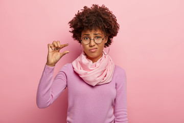 Upset curly haired woman shapes little gesture, demonstrates something very tiny, wears optical glasses, casual jumper and scarf, measures small size, isolated over pink background. Not very much