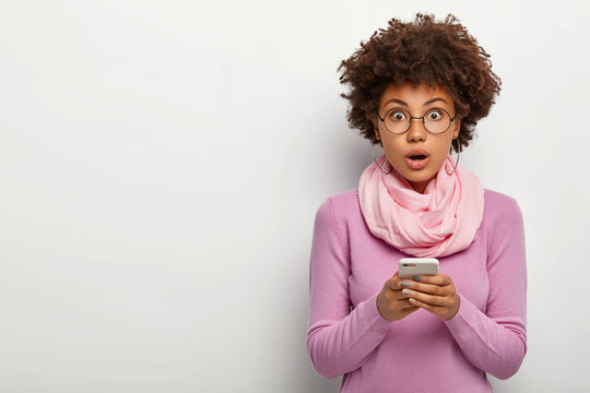 Photo of surprised curly haired woman wears glasses, holds mobile phone, gets message, looks with shocked expression at camera, wears round eyewear, casual outfit, poses indoor. People, lifestyle