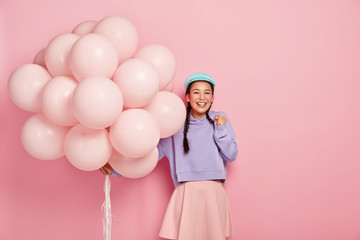 Photo of happy Asian girl clenches fist with joy, cant wait for special moment, gets congratulation from friends on her birthday, carries big bunch of balloons, dressed in fahionable clothes