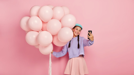 Half length shot of smiling charming dark haired Korean woman takes selfie portrait on modern cellular, poses with bunch of balloons, has fun on party, celebrates special occasion, has playful mood