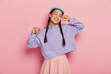 Carefree cheerful millennial Asian woman keeps hands up, dances against pink background, has eyes closed, gets pleasure from favourite music, being well dressed, has good mood, positive emotions