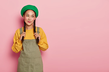Thoughtful Asian lady daydreams about something, touches braids, dressed in stylish outfit, has spare time, stands against pink background, copy space on right side for your promotional content