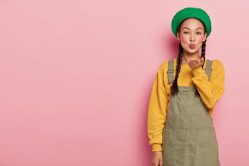 Beautiful young female with Asian appearance, blows air kiss, keeps lips rounded, wears makeup, expresses love to someone, wears fashionable clothes, isolated on pink studio wall. Body language