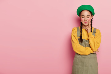 Pleasant looking satisfied woman with delighted expression, keeps index finger on cheek, eyes shut, models against pink background, gets pleasure from hearing good heartwarming words from guy