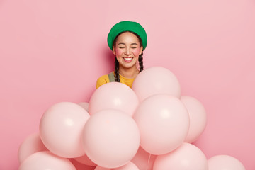 Obraz na płótnie Canvas Smiling woman with cheerful expression, keeps eyes closed from pleasure, wears green beret, stands with inflated helium balloons, has two pigtails, gets congratulation with birthday or anniversary