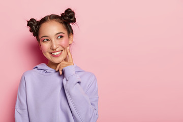 Studio shot of glad lovely woman with smile, keeps index finger on cheek, looks aside, has two...