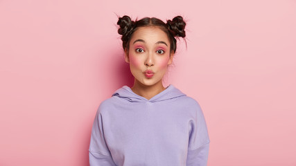 Facial expressions concept. Pretty young Japanese woman with funny look, keeps lips rounded, has two hair buns, wears casual purple hoody, isolated over pink background, wants to kiss someone