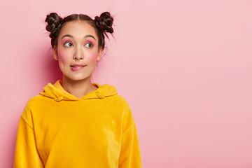 Portrait of thoughtful young woman with two hair buns, bites lips, being deep in thoughts, tries to find solution in mind, dressed in yellow jumper, has childish hairstyle, poses against pink wall