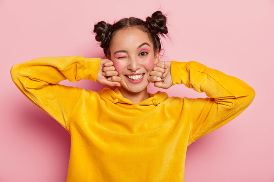 Image of lovely young Asian woman with happy face expression, blinks eye and smiles positively, has fun indoor, two hair knots, dressed in yellow velvet hoody, isolated over pink background.