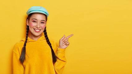 Friendly looking cheerful Asian woman points fore finger aside, recommends cool advertisement, has two braids, wears stylish cap and yellow sweatshirt, sure you will like it, suggests buying something